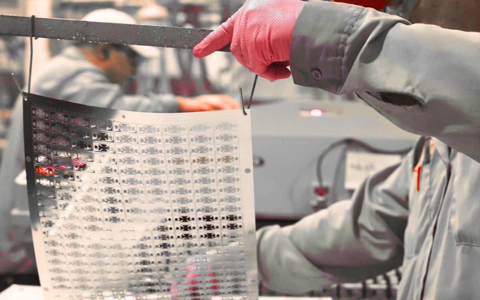 Worker wearing gloves holding up a thin precision-etched metal component sheet in a manufacturing facility.